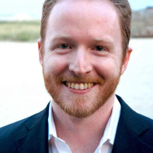 wpa_people_page_reb_rebele Headshot of a person with short hair and a beard, smiling, wearing a suit jacket and white shirt, with an outdoor background.
