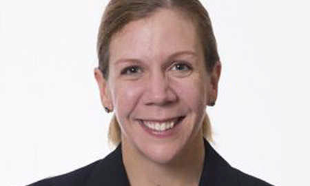 Headshot of a person smiling, wearing a dark blazer with light-colored hair pulled back.