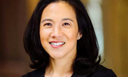 Headshot of a smiling person with shoulder-length dark hair and a dark top, set against a blurred background.