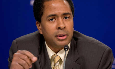 A person speaking at a press conference, wearing a suit and tie, against a blue background.