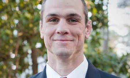 Headshot of a person wearing a suit and tie, smiling outdoors with trees in the background.