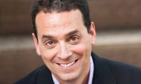 A headshot of a smiling person wearing a suit jacket and shirt, with short, curly hair in a casual setting.