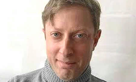 A headshot of a person with light hair wearing a gray turtleneck sweater against a plain background.