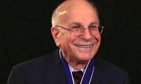 Headshot of a smiling person with glasses and a medal, wearing a suit and tie, against a dark background.