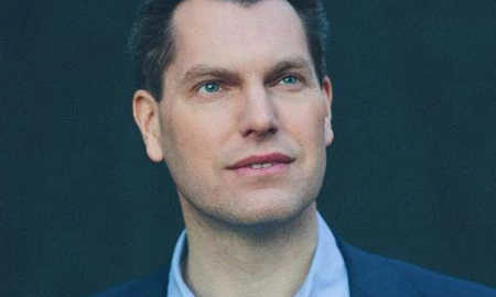 Headshot of a person with short hair, wearing a collared shirt and jacket, looking slightly upward against a dark background.