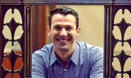 A person with short dark hair and a wide smile, wearing a blue shirt, framed by ornate wooden carvings.