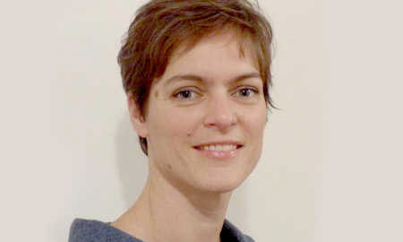 A headshot of a person with short hair, wearing a gray top, smiling against a plain background.