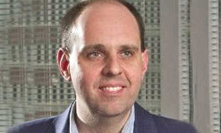 Headshot of a person with a neutral expression, wearing a suit, with short hair, in an indoor setting featuring office blinds.