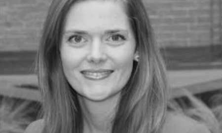 Headshot of a person with long hair, smiling, in front of a blurred brick wall background, in black and white.