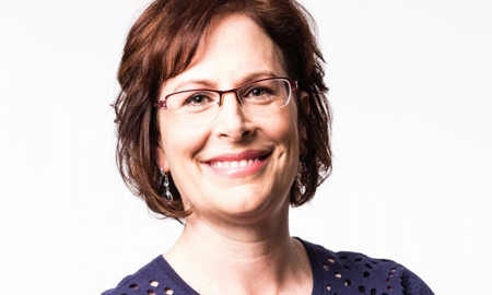 A headshot of a person with short, brown hair and glasses, smiling, wearing a dark blue top.