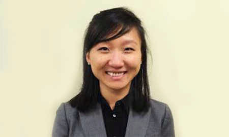 A headshot of a person with long dark hair, wearing a gray blazer and a black shirt, smiling at the camera.