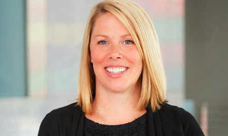 A headshot of a person with shoulder-length blond hair, smiling at the camera, wearing a black top. The background is softly blurred.