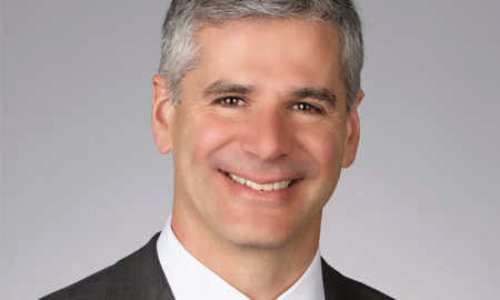 Headshot of a smiling person with short gray hair wearing a dark suit and white shirt against a plain background.