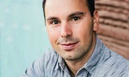 Headshot of a person with short dark hair wearing a gray button-up shirt, smiling slightly, with a blurred background.