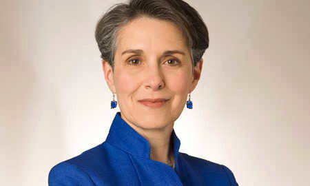 A person wearing a blue jacket and blue earrings, smiling slightly, with short gray hair. The background is a plain, light color.