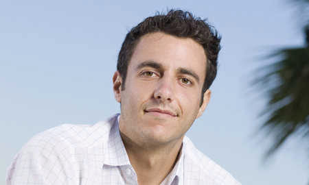 A headshot of a person with short dark hair, wearing a light-colored shirt, with a clear blue sky and a hint of palm tree in the background.