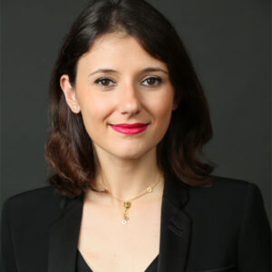federica_resized Headshot of a person with shoulder-length dark hair, wearing a black blazer and a necklace, set against a dark background.