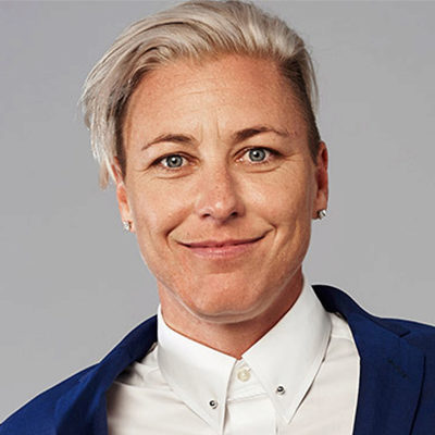 A person with short, light-colored hair, wearing a blue suit and a white shirt, smiling, with a neutral background.