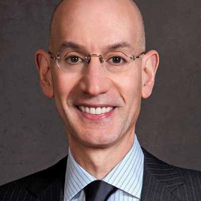 Headshot of a person wearing glasses, a suit, and a tie, smiling against a neutral background.