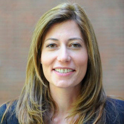 Headshot of a person with long, light brown hair, smiling, against a blurred background.