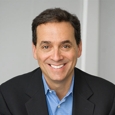 Headshot of a person with short dark hair, wearing a blue shirt and dark suit jacket, smiling at the camera.