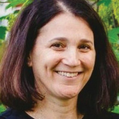 Headshot of a person with shoulder-length dark brown hair, smiling outdoors with green foliage in the background.