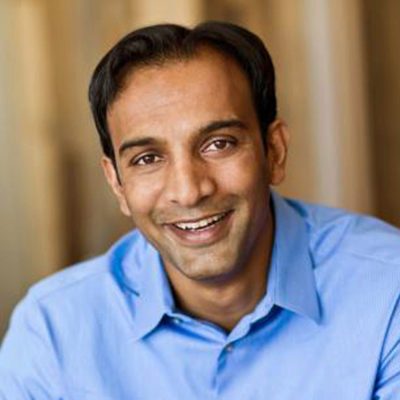 A person in a blue shirt smiling directly at the camera, set against a blurred background.