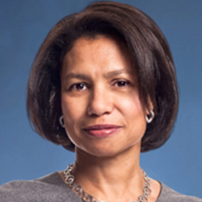 Headshot of a person with short, dark hair, wearing a gray top and a necklace, set against a blue background.