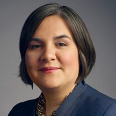 Headshot of a person with short dark hair, wearing a dark blazer and a necklace, against a neutral background.