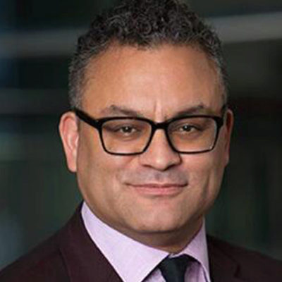 Headshot of a person with short curly hair, wearing glasses, a suit, and a tie, smiling slightly. Background is blurred and dark.