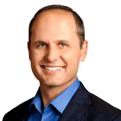 A headshot of a person smiling, wearing a blue shirt and dark jacket against a plain white background.