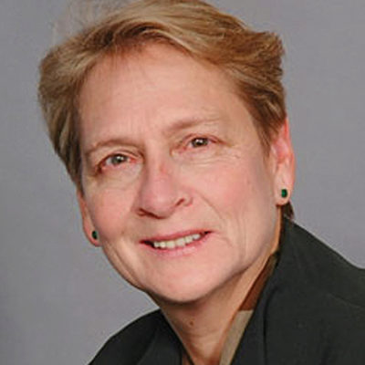 Headshot of a person with short, light brown hair wearing a dark jacket, set against a plain gray background.