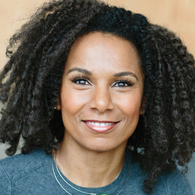 Headshot of a person with curly hair, smiling, wearing a gray top, in front of a neutral background.