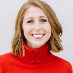 A person with shoulder-length hair is smiling, wearing a bright red turtleneck sweater. The background is plain and light-colored. Headshot.