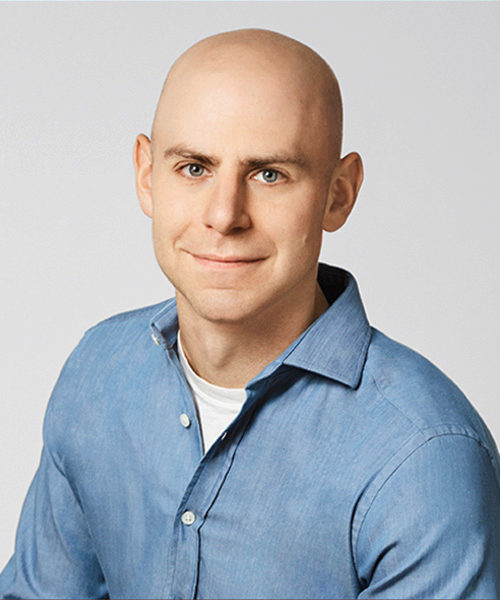 Headshot of a person with a bald head, wearing a blue button-up shirt and a white T-shirt underneath, smiling slightly, with a neutral background.