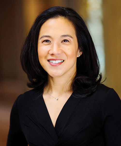 Headshot of a person with shoulder-length black hair, smiling, in a black outfit with a necklace, set against a softly blurred background.