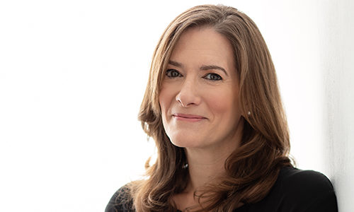 Headshot of a person with long brown hair, smiling softly, dressed in a dark top, against a light background.