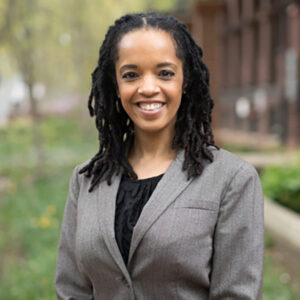 A person with long braided hair is smiling, wearing a gray suit jacket, and standing outdoors with greenery and buildings in the background.