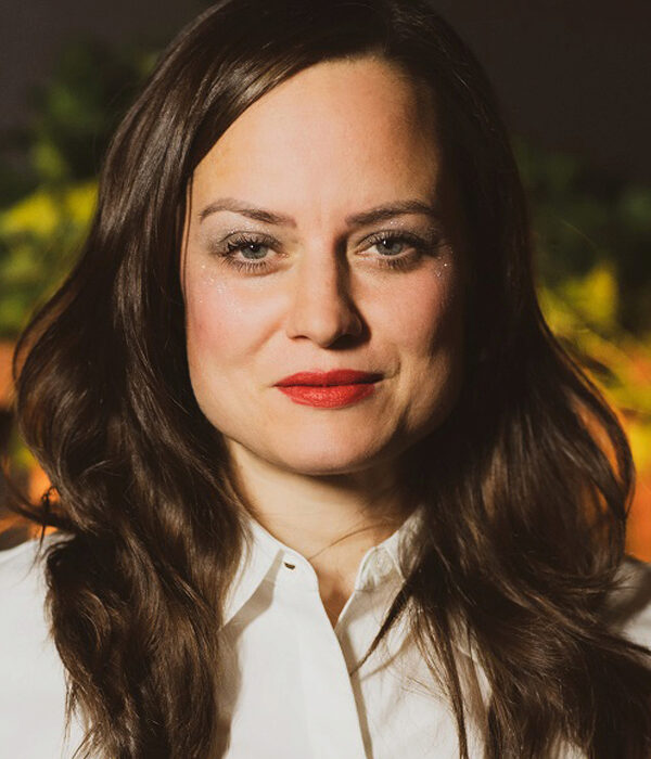 Headshot of a person with long brown hair, wearing a white shirt, with a neutral expression. The background is softly blurred with greenery.