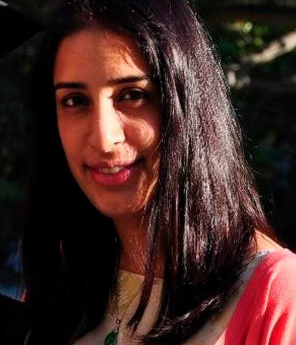 Headshot of a person with long dark hair, smiling, wearing a light-colored top and a pink cardigan, outdoors in natural light.