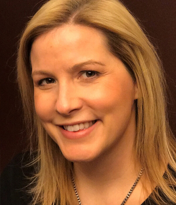 Headshot of a person with blonde hair and a slight smile, wearing a necklace, against a dark background.