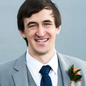 Louis Hickman rev A headshot of a person in a gray suit and navy tie, smiling. They have a boutonniere with orange flowers on the lapel.