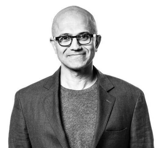 A black and white headshot of a person wearing glasses and a suit jacket, smiling slightly, with a plain background.