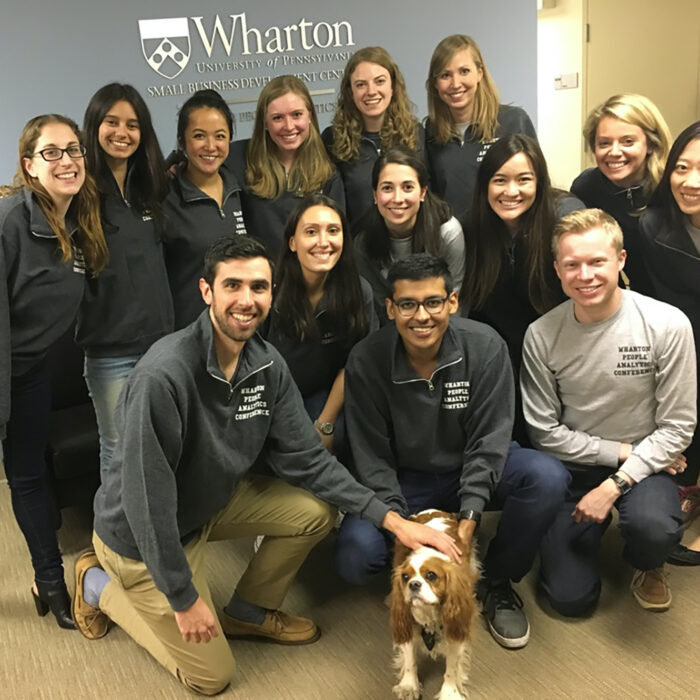 A group of people posing indoors, wearing matching Wharton-branded clothing, with a dog sitting in front of them. The background features a Wharton University sign.