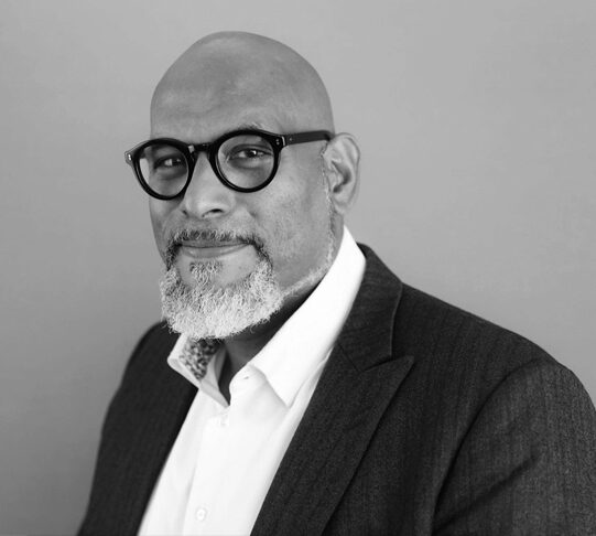 A black and white headshot of a person wearing glasses, a suit jacket, and a collared shirt, set against a plain background.