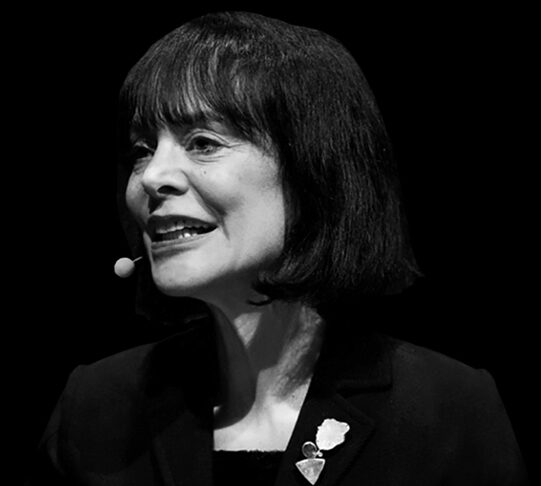 A black and white headshot of a person speaking into a microphone, wearing a suit with pins on the lapel.