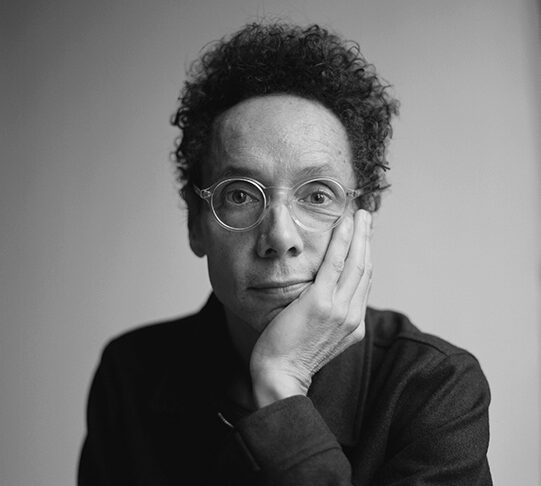 Black and white headshot of a person with curly hair and glasses, resting their face on a hand, wearing a collared shirt or jacket.