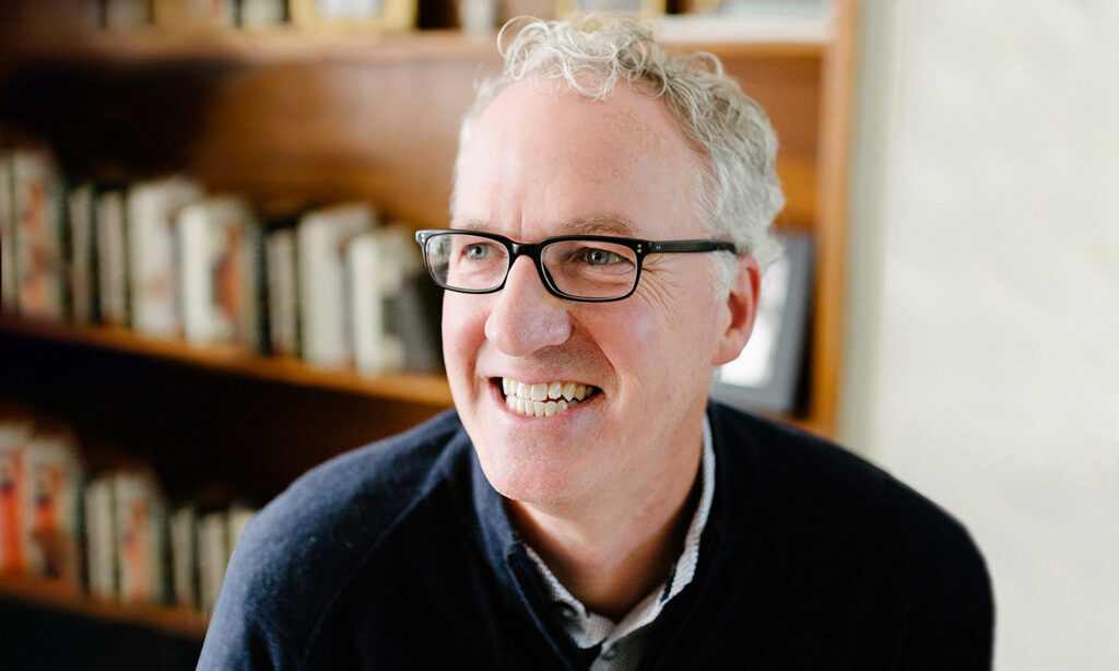 Smiling person with glasses and curly hair, wearing a dark sweater, in a room with bookshelves in the background.