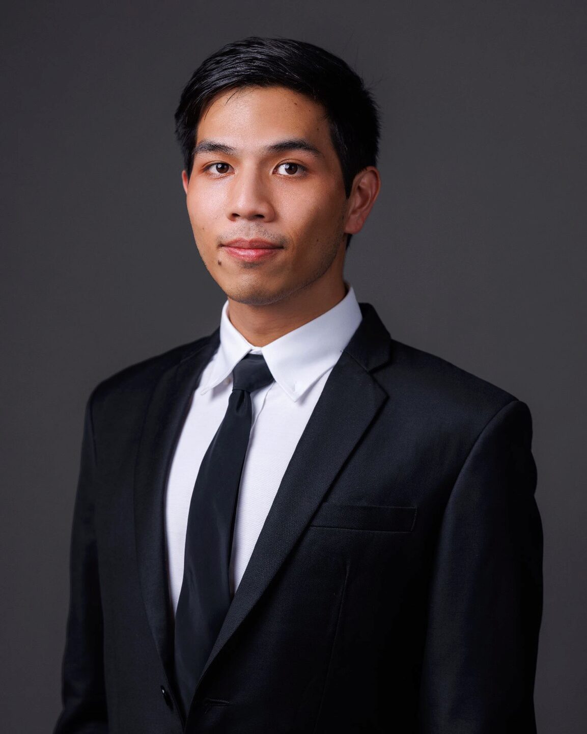 A headshot of a person wearing a black suit and tie with a white shirt, set against a dark background.