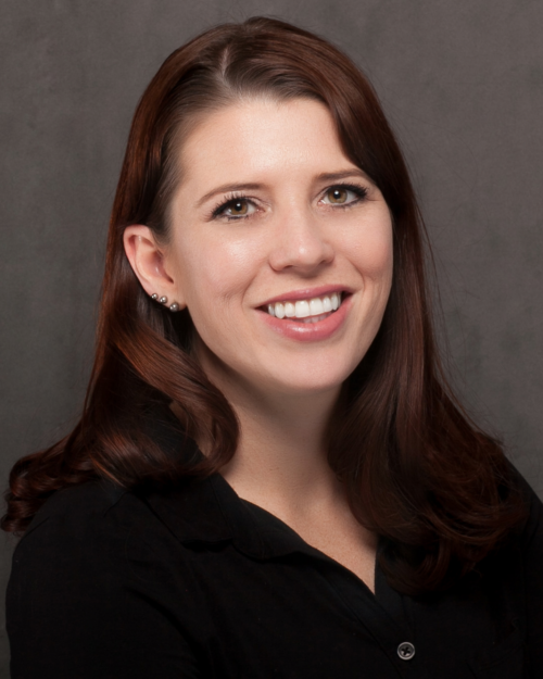 Headshot of a person with long brown hair, wearing a black shirt, smiling against a gray background.